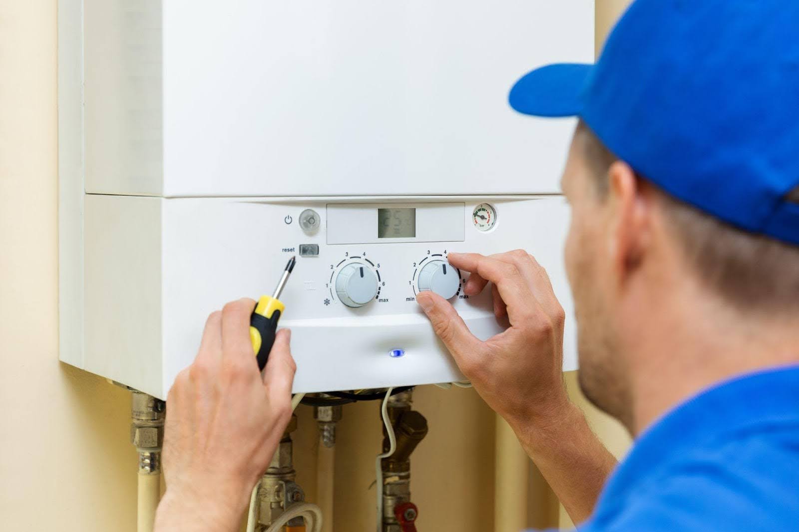A man wearing a blue shirt and hat repairs a gas boiler focused on his task in a domestic setting