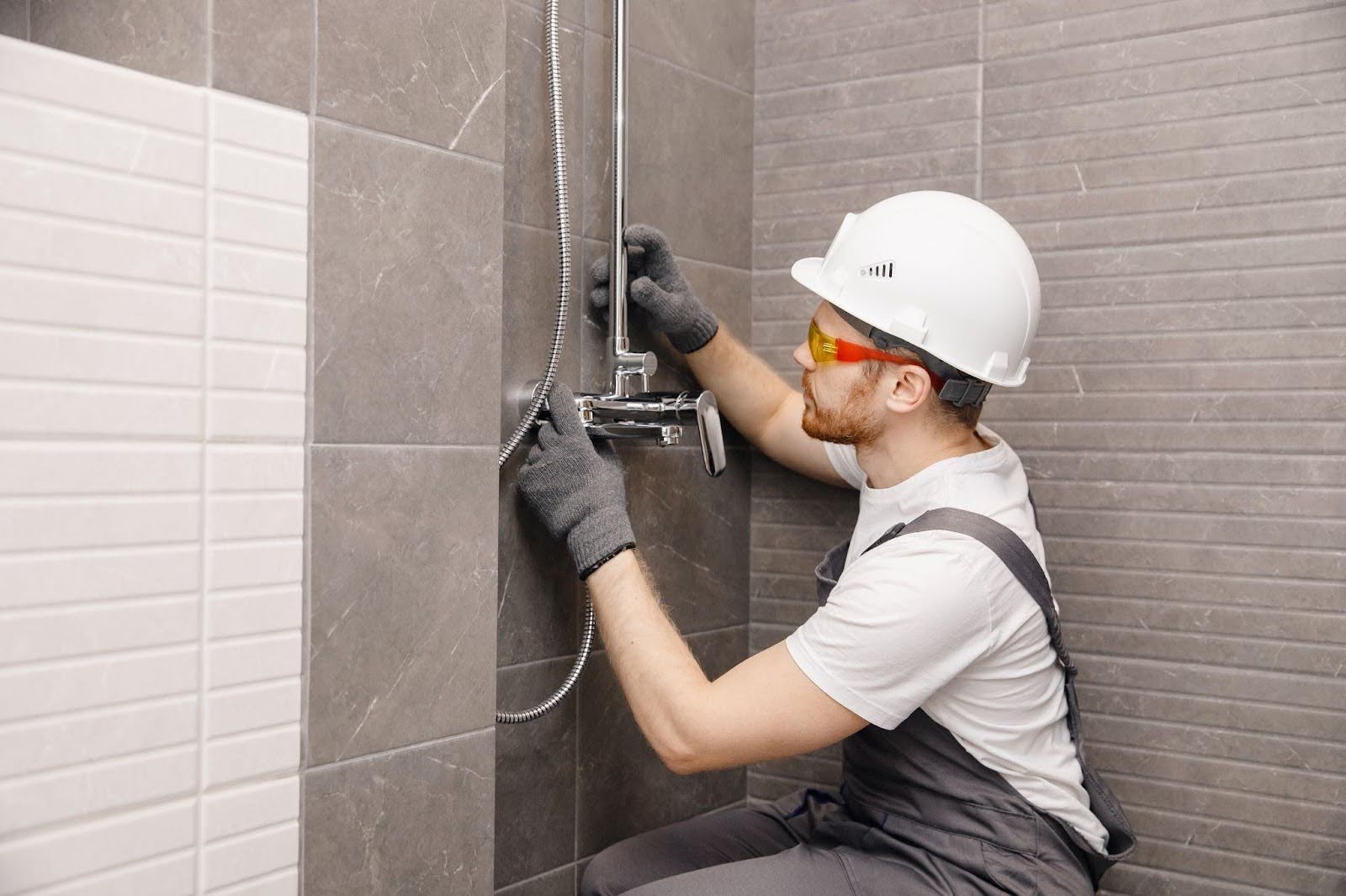 A man wearing a hard hat and safety glasses repairs a shower head in a bathroom setting
