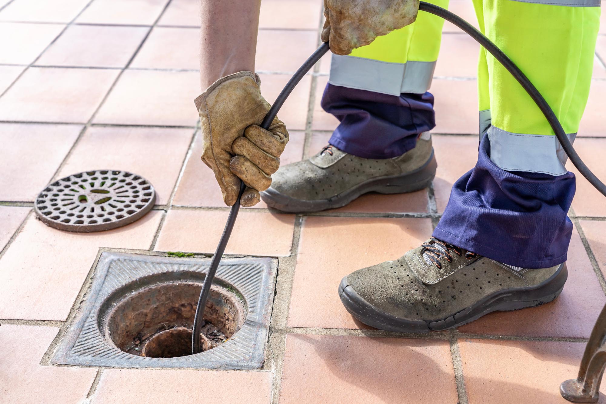A worker in gloves uses a drain cleaning tool to unclog a sewer drain on a tiled surface.