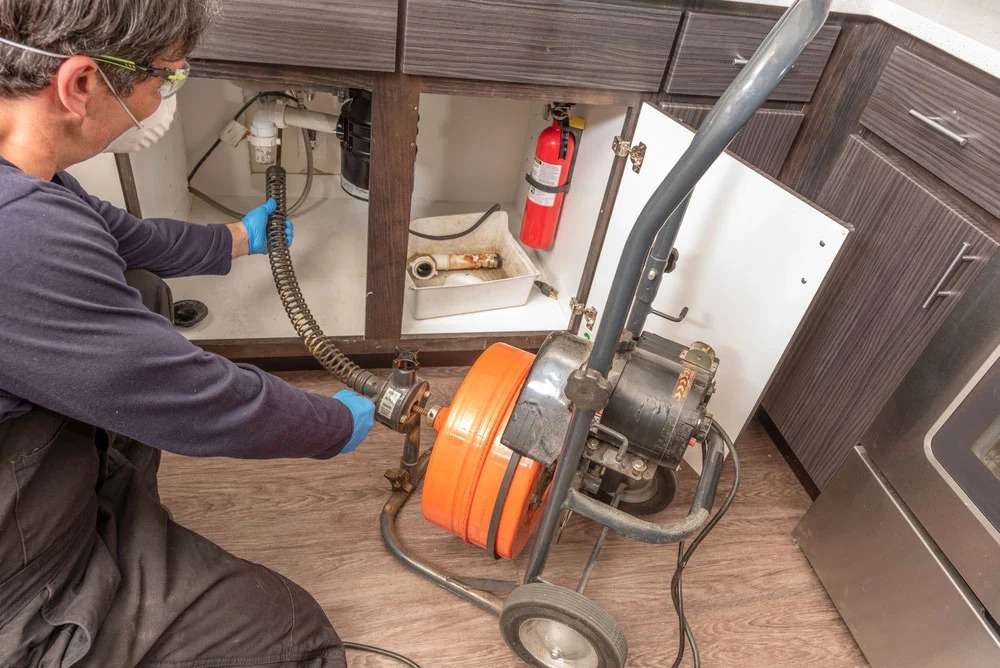 A plumber wearing a mask and gloves uses a professional drain cleaning machine to clear a clog under a kitchen sink with dark wood cabinets.