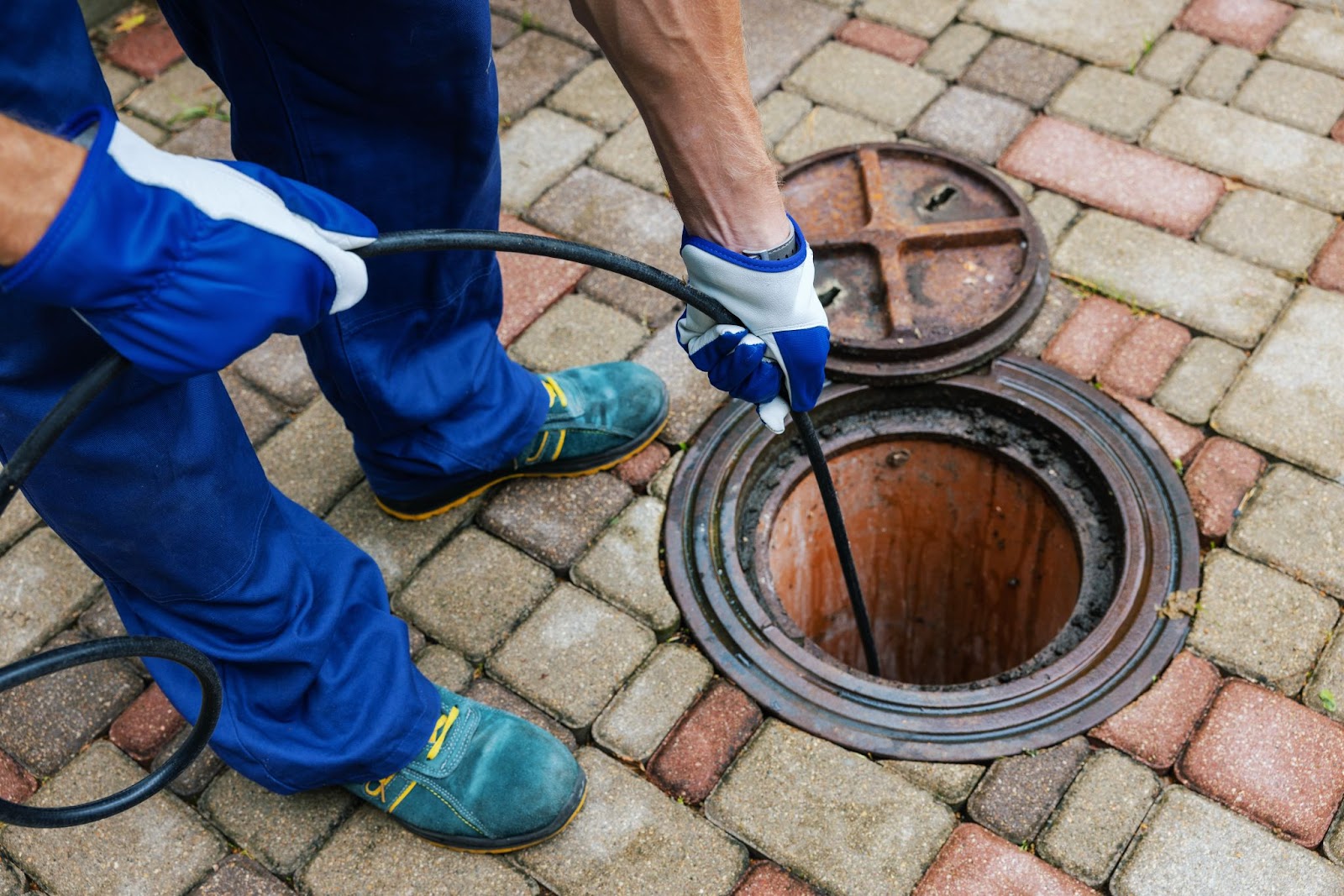 A worker in blue uniform and protective gloves inserts a long black cable into an open sewer manhole on a paved brick walkway for inspection.