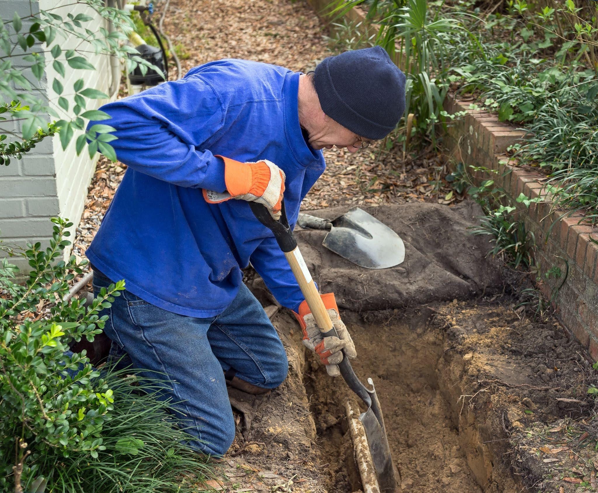 A man wearing a blue shirt and beanie kneels in a garden, using a shovel to dig a narrow trench in the soil next to a brick retaining wall.