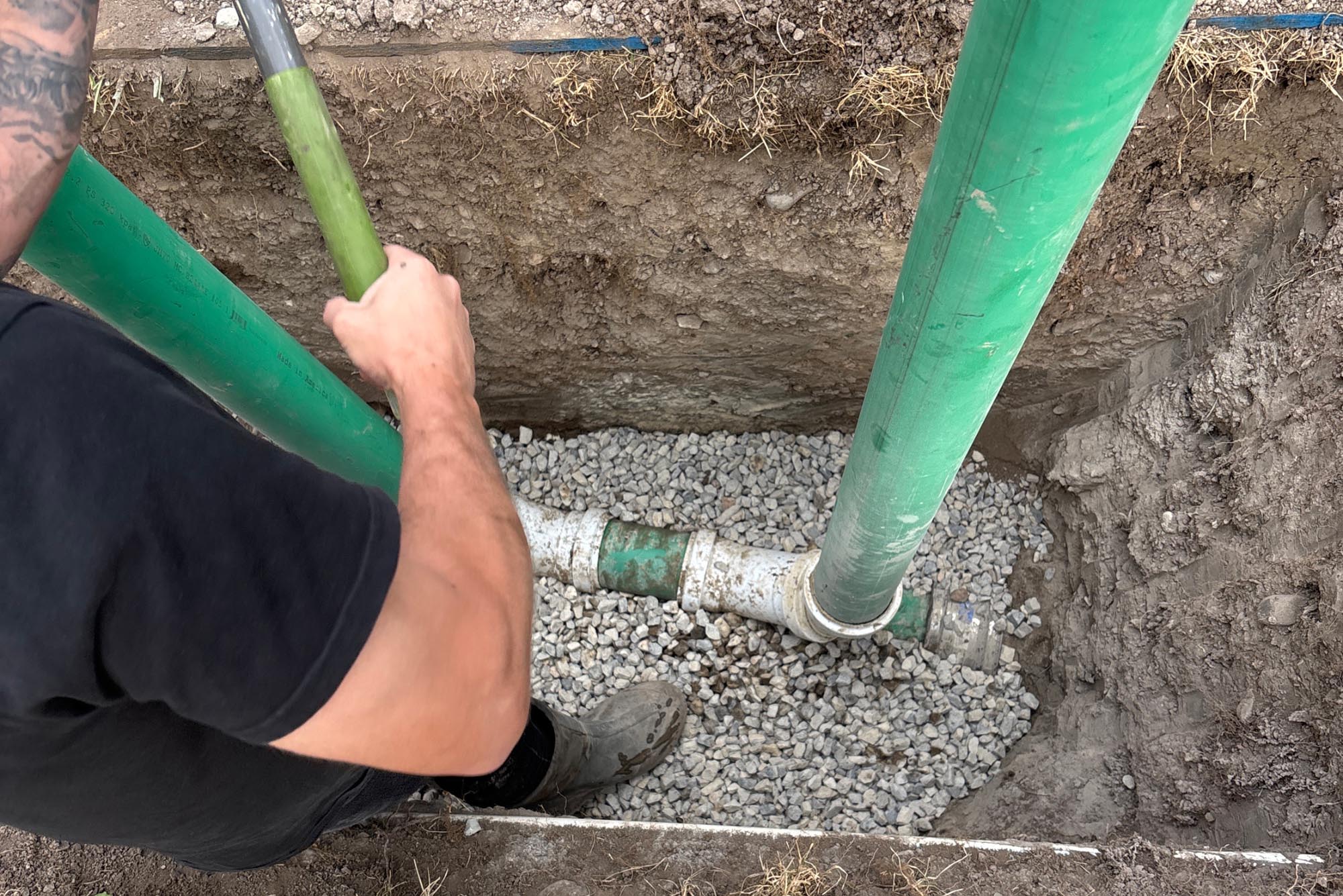 A construction worker uses a shovel to spread gravel around green PVC pipes installed in a narrow trench during a plumbing or drainage project.
