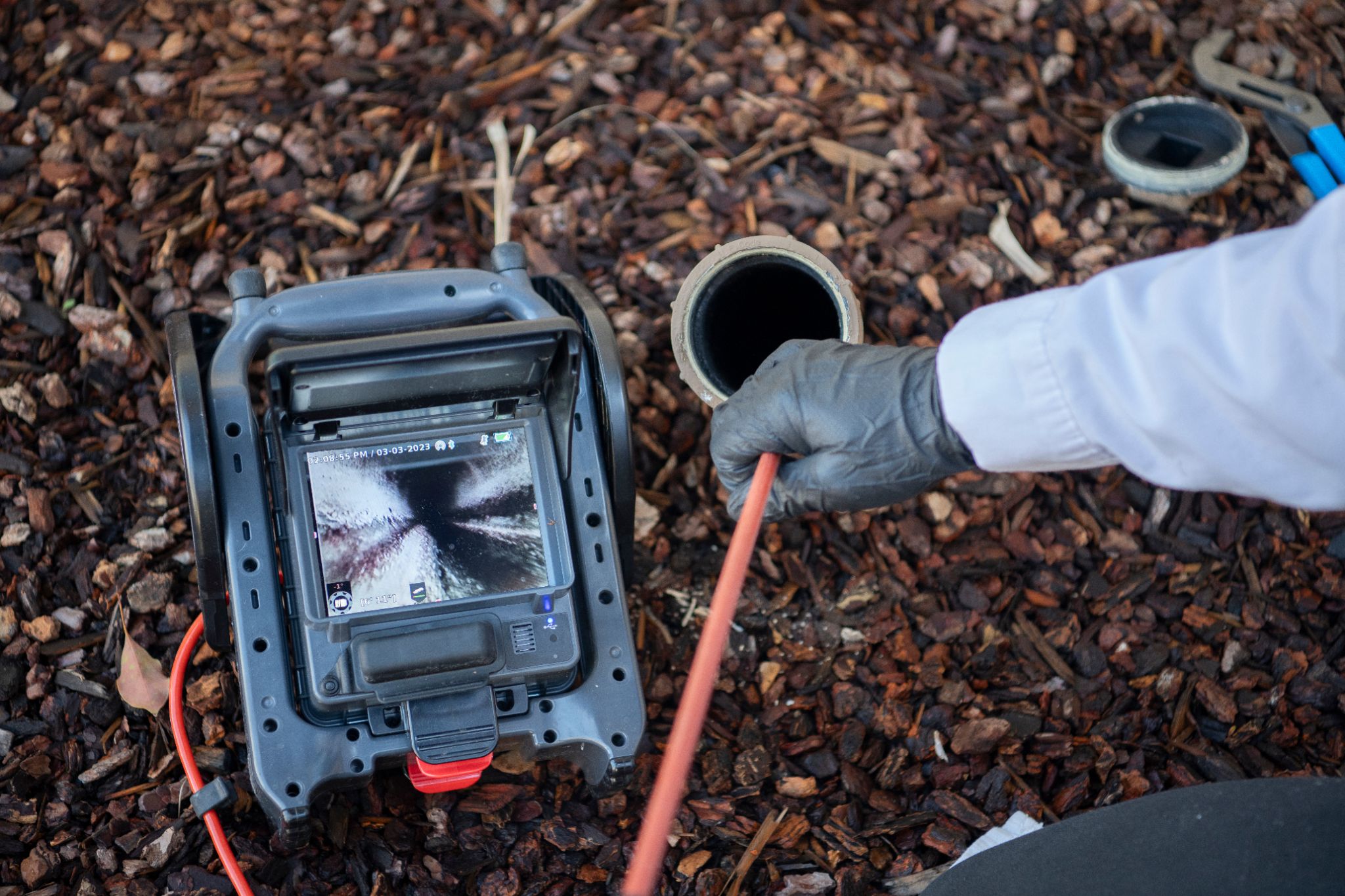 Plumber feeding an inspection camera into an outdoor drain pipe next to a video monitor.