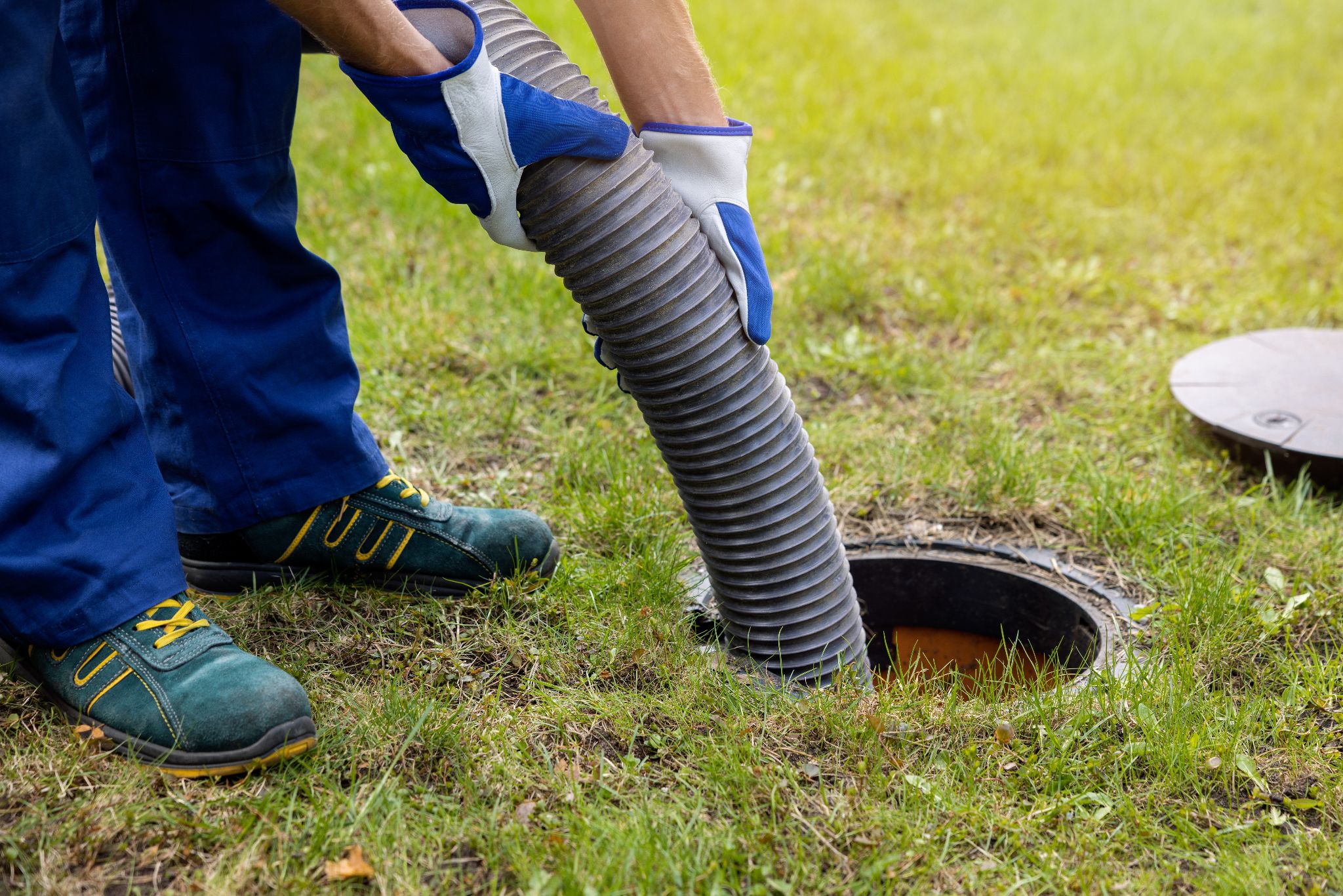 Worker inserting a suction hose into an outdoor septic tank opening on a lawn.