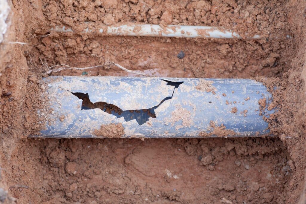 A damaged blue underground water pipe is exposed in a soil trench with a large jagged crack