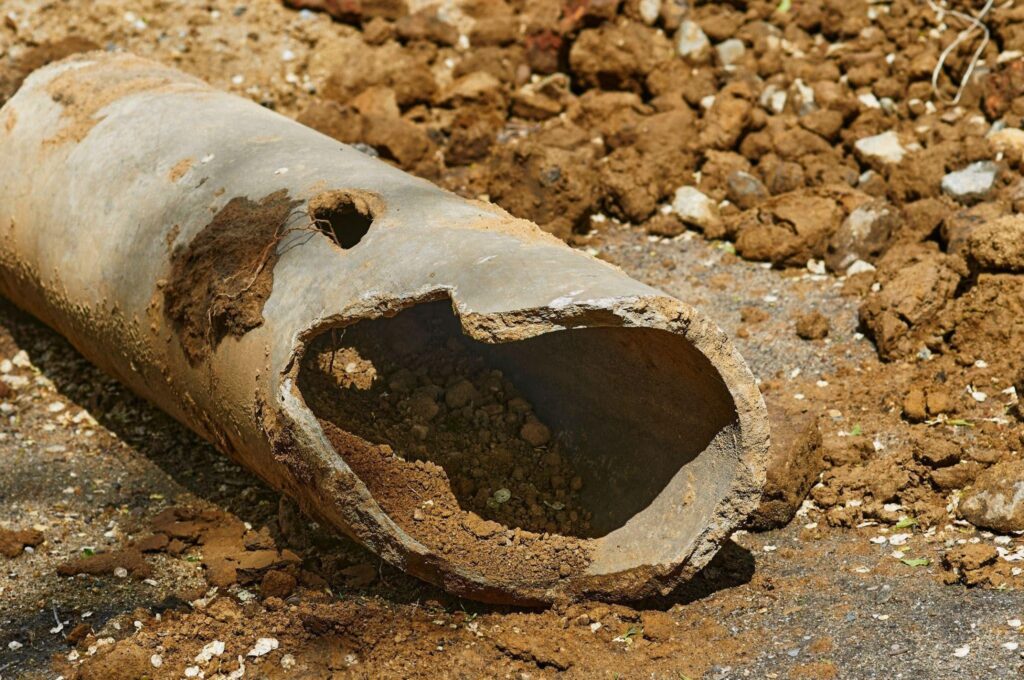 A weathered rusted metal pipe with a jagged broken opening lies amid dry soil rocks and debris