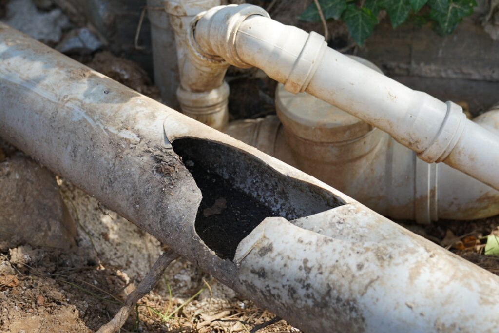 A close-up view of a damaged, broken white PVC drainage pipe lying on the ground, revealing dark soil inside, with other plumbing pipes nearby.