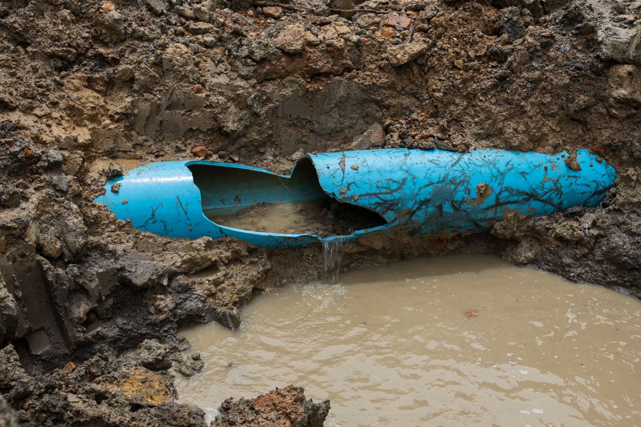 A broken blue plastic pipe lies in a muddy trench, with water leaking from the damaged section into a pool of murky water in the excavated ground.